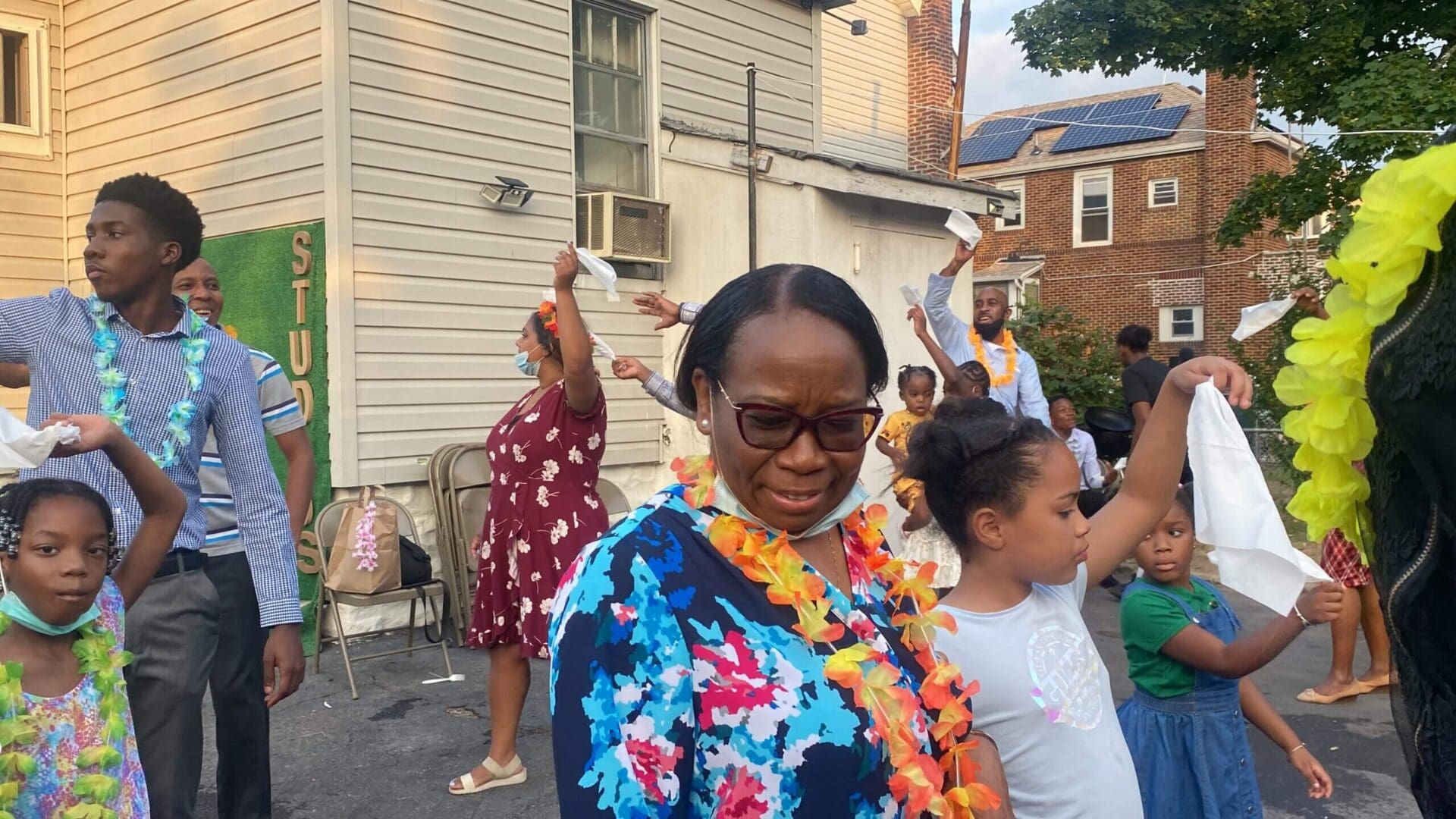 A joyful woman in a floral dress and lei at an outdoor gathering.