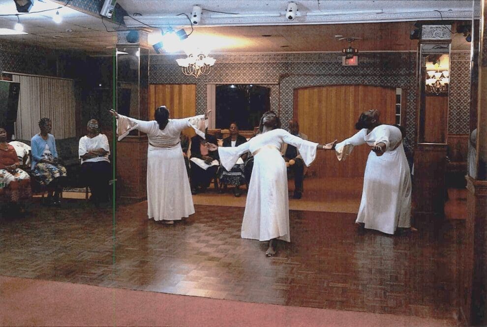 Three women in white dresses dancing indoors in front of seated audience.